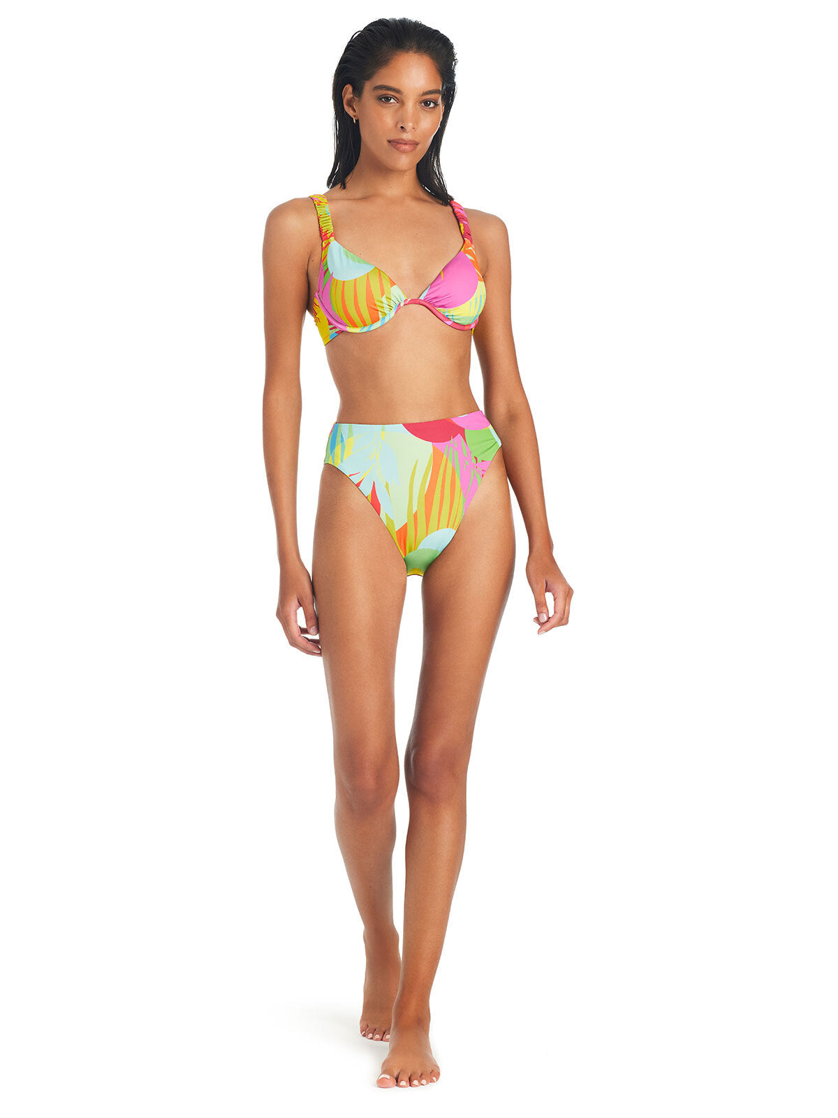 A woman stands in a Sanctuary Clothing Palm Hottie High Rise Bikini Bottom Multi Palm Hottie, featuring a supportive top with a vibrant tropical pattern. Her long, dark wet hair and confident expression complement her relaxed pose with hands by her sides. The white background emphasizes her stylish swimwear.
