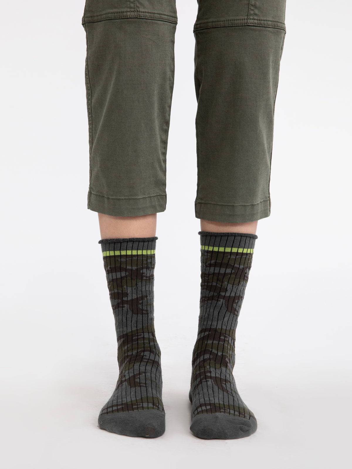 The image shows a person wearing green pants that end slightly below the knees, along with a pair of Heritage Camo Socks by Sanctuary Clothing, which feature a mix of dark and light green and gray colors. The person is standing on a white background.