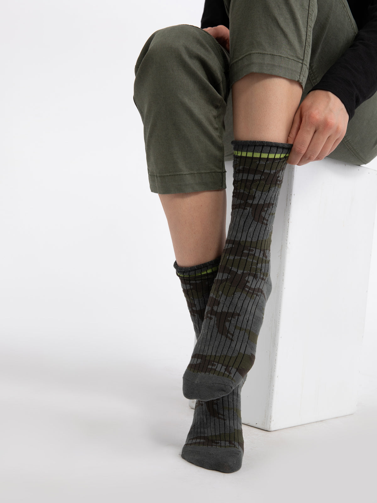 A seated person wearing green pants reveals their lower legs adorned with Sanctuary Clothing's Heritage Camo Socks. Their right foot is relaxed on the ground while their left leg crosses over it, resting on the opposite knee. The background is plain white.