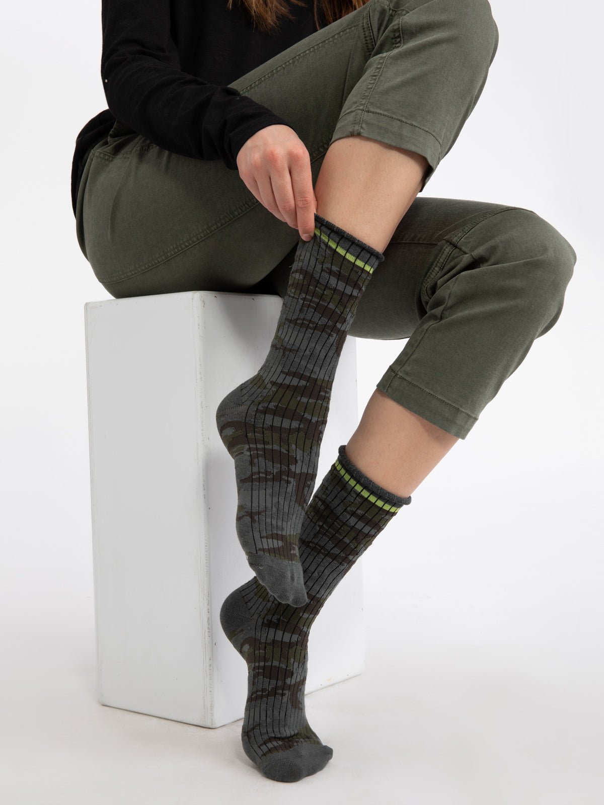 A person seated on a white block, adjusting one of their two Sanctuary Clothing Heritage Camo Socks. They are wearing olive green pants and a black top, with their right leg crossed over the left knee.