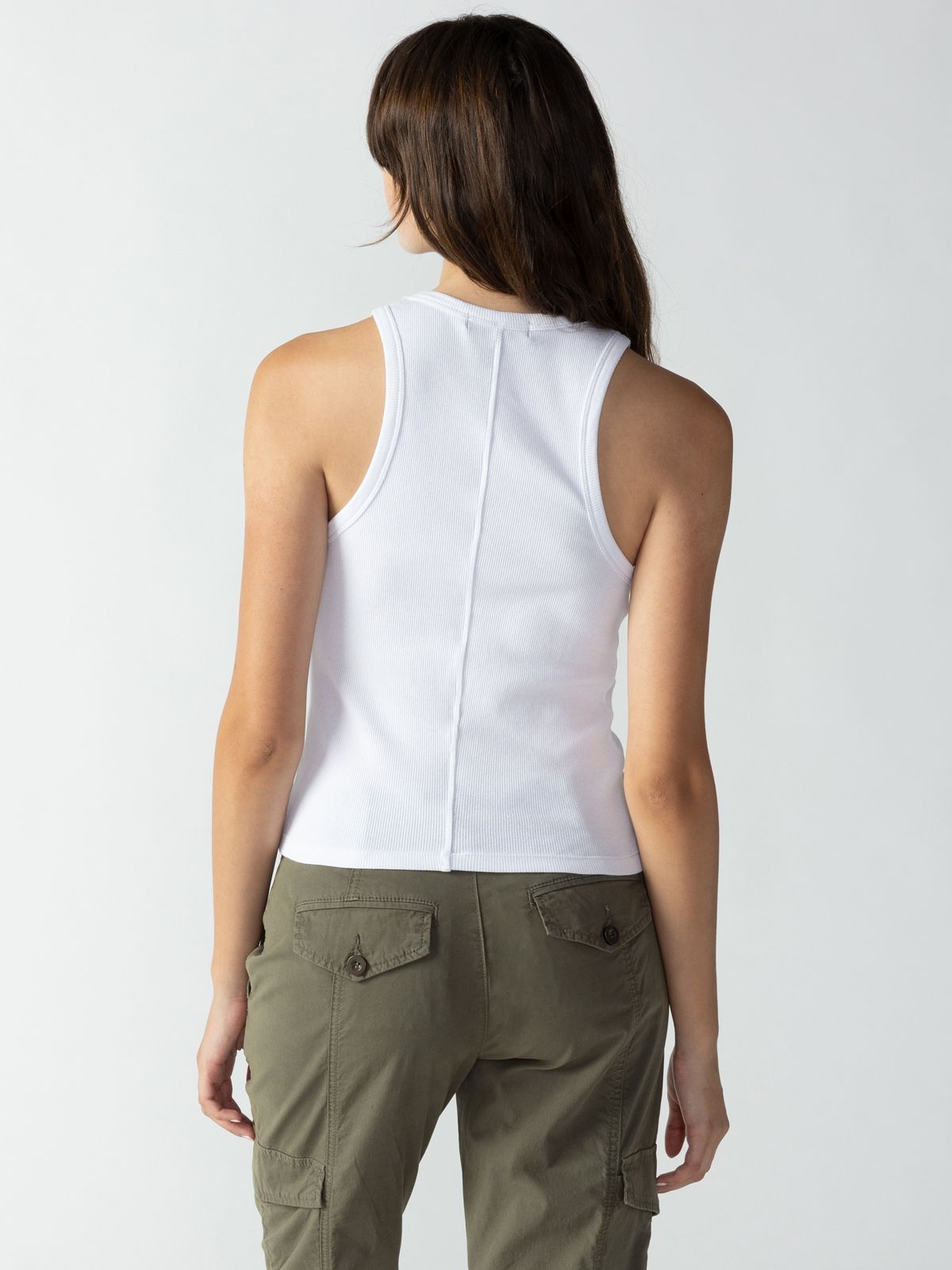 A person with long brown hair is seen from the back, wearing a sleeveless Ribbed Tank Top in white by Sanctuary Clothing and a pair of olive green cargo pants. The background is plain and light-colored.