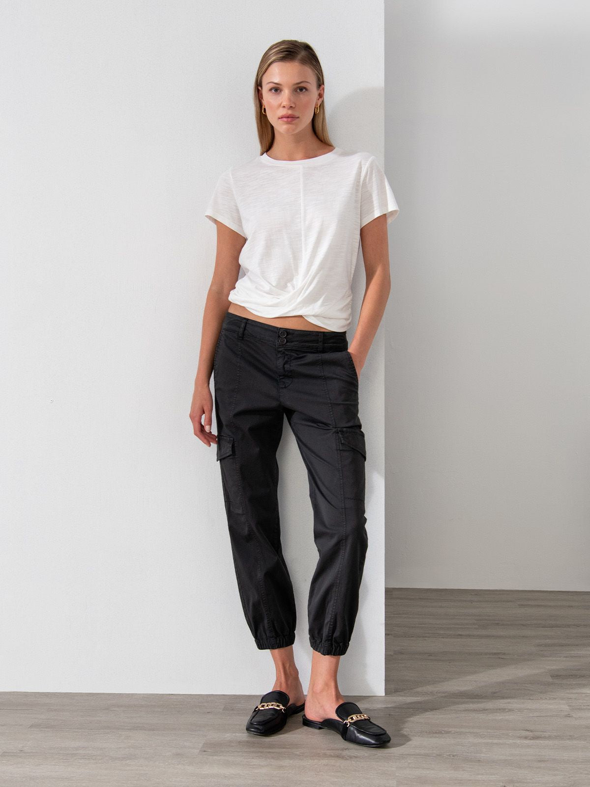 A woman stands indoors against a white wall, wearing the Sanctuary Clothing Highstreet Tee in white, black cargo pants, and black loafers. She has one hand in her pocket and looks straight at the camera with a neutral expression. The floor is wooden and there are no decorations visible.