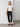 A woman stands indoors against a white wall, wearing the Sanctuary Clothing Highstreet Tee in white, black cargo pants, and black loafers. She has one hand in her pocket and looks straight at the camera with a neutral expression. The floor is wooden and there are no decorations visible.