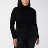 A person with medium-length dark hair is wearing the Essential Turtleneck Black from the Sanctuary Clothing Inclusive Collection and black jeans. They stand against a plain white background, looking directly at the camera with a confident and composed expression.