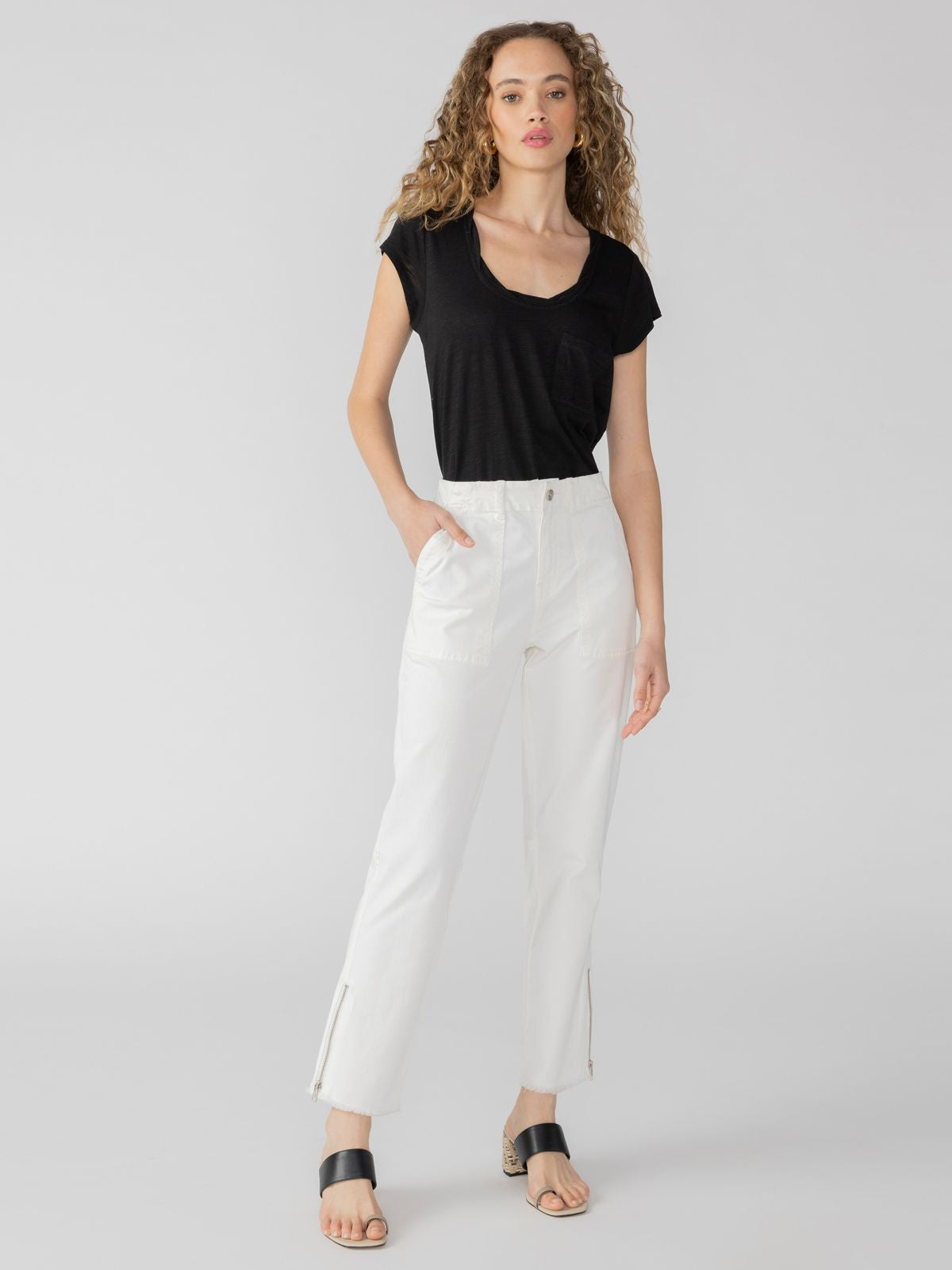 A person with curly hair stands against a plain background. They are wearing the Traveler Twist Tee in black from Sanctuary Clothing, white high-waisted pants with side pockets and ankle zippers, and black sandals. One hand is in a pocket while the other rests at their side.