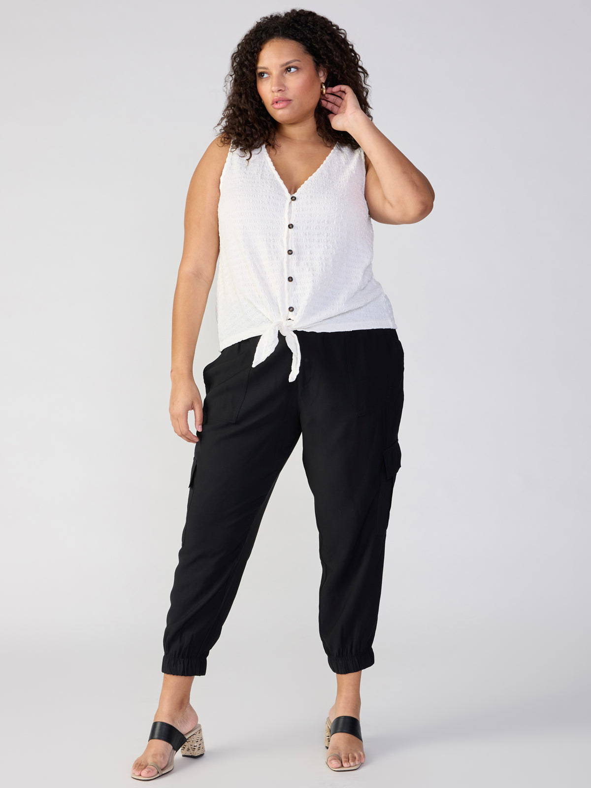 A woman with curly hair is standing against a plain background, wearing a white sleeveless top with a front tie and The Harmony Black jogger pants by Sanctuary Clothing. She is holding her left hand near her face and is dressed in open-toe sandals.