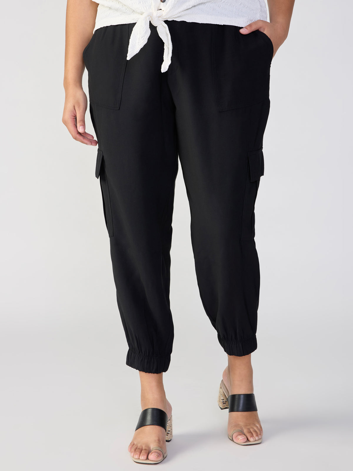 A person is wearing The Harmony Black cargo pants with elasticated cuffs from Sanctuary Clothing, standing with one hand in their pocket. They are also wearing black slide sandals featuring block heels and a white top tied at the waist. The background is plain and neutral.