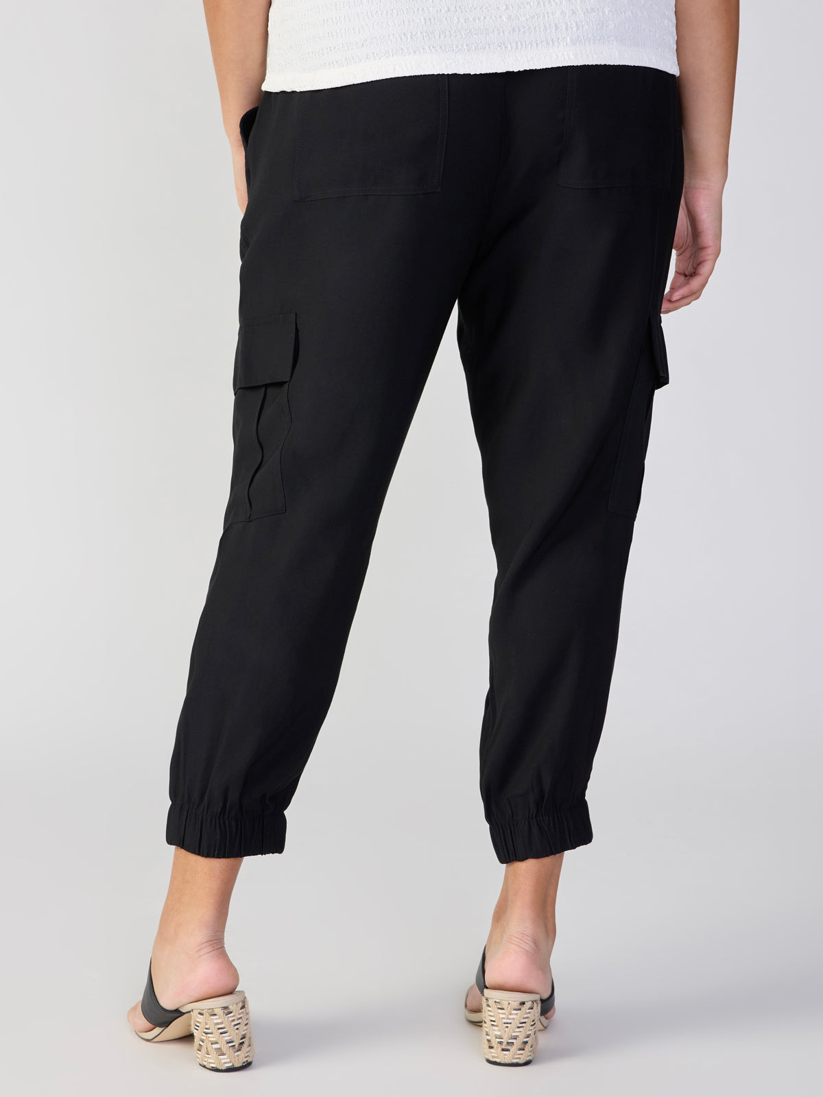 A person is wearing The Harmony Black cargo pants from Sanctuary Clothing, featuring elastic cuffs and cargo pockets on the side, paired with a white top. They are also sporting open-heel shoes with block heels. The photo is taken from the back, highlighting the details of the pants.