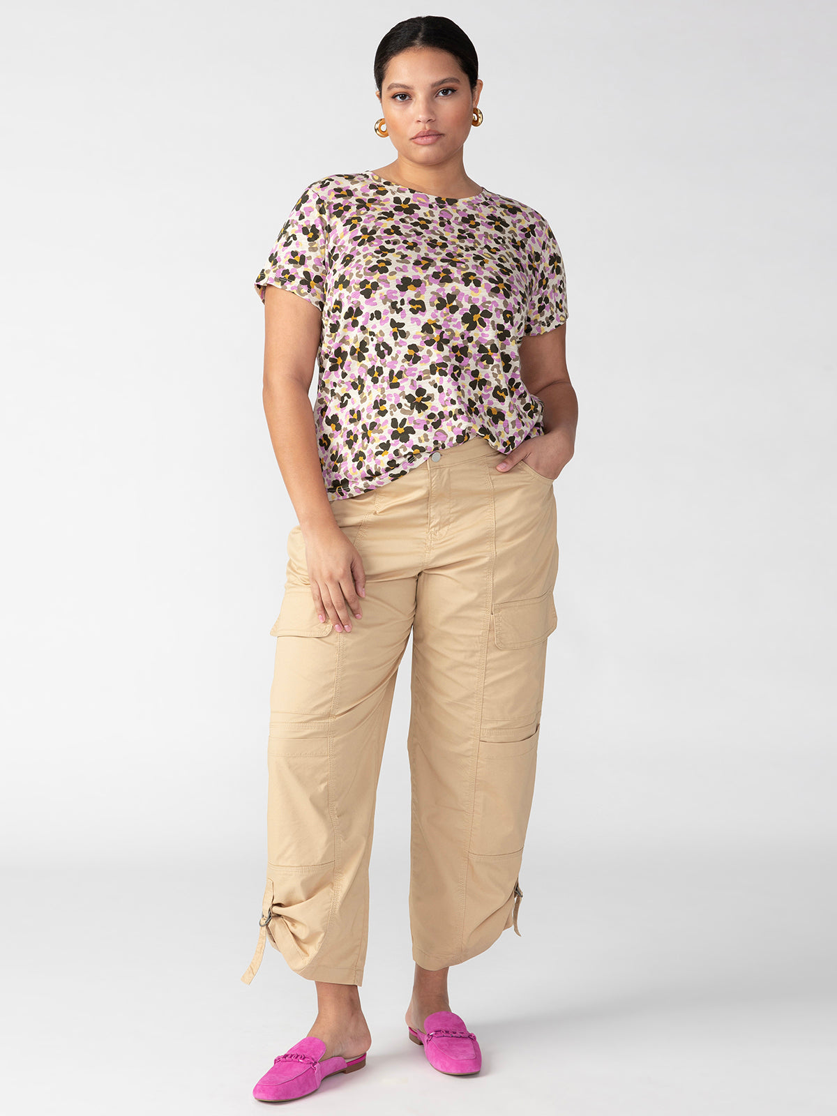 A woman stands against a plain background wearing a floral-patterned short-sleeve shirt, Sanctuary Clothing's Cali Cargo True Khaki Inclusive Collection beige cargo pants, and pink loafers. She has her hand in one of the pockets and is looking at the camera with a neutral expression. She also has short dark hair and is wearing hoop earrings.