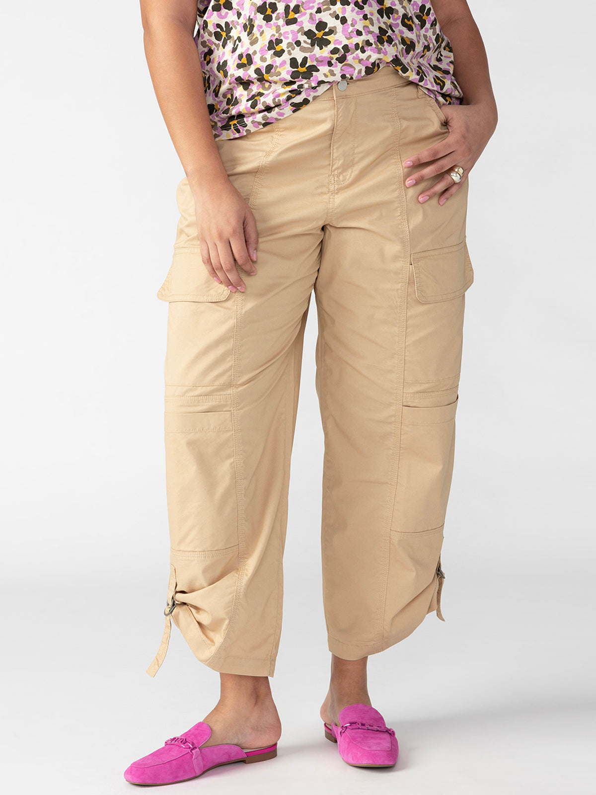 A person stands against a plain white background, wearing the Cali Cargo True Khaki Inclusive Collection pants by Sanctuary Clothing. They pair these with a colorful floral-patterned top, bright pink slip-on shoes, and a ring on their left hand.