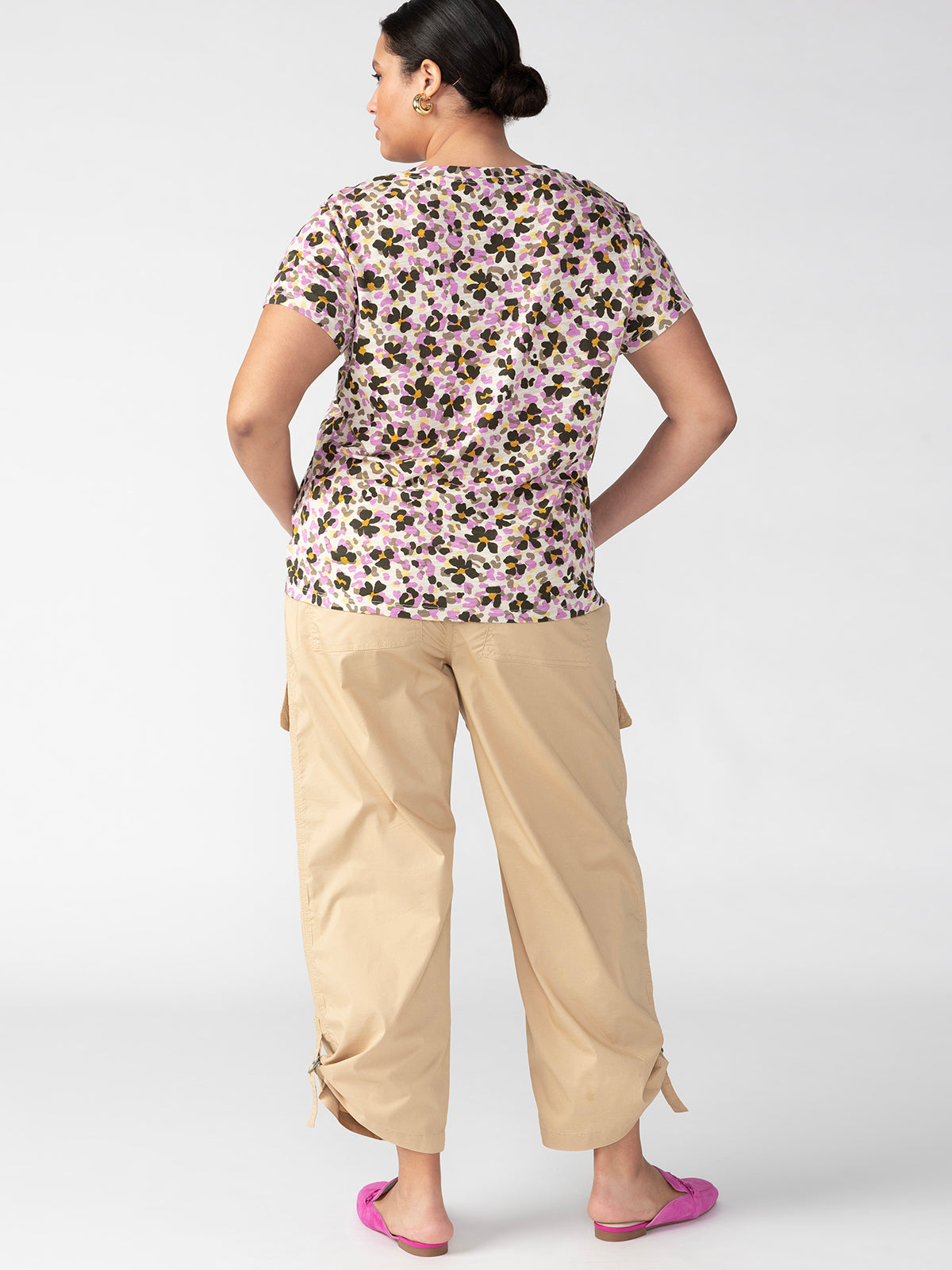 A person is standing with their back to the camera, wearing a floral short-sleeve shirt, Cali Cargo True Khaki Inclusive Collection pants from Sanctuary Clothing, and pink slip-on shoes. The background is plain white.