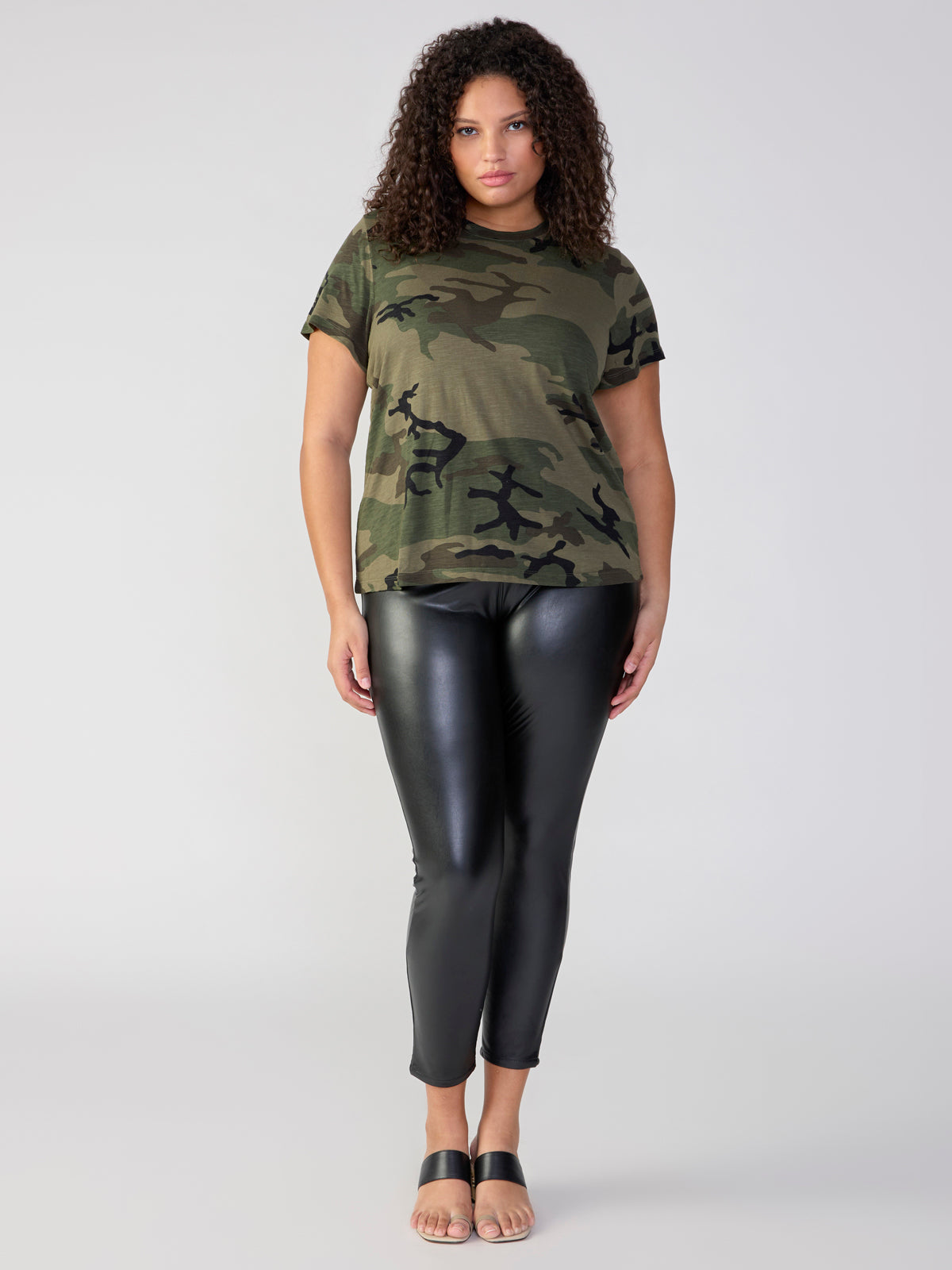 A person with curly hair stands facing the camera. They are wearing a short-sleeve green camouflage T-shirt, Sanctuary Clothing's black Runway Legging from the Inclusive Collection, and open-toed black sandals. The background is plain and light-colored.