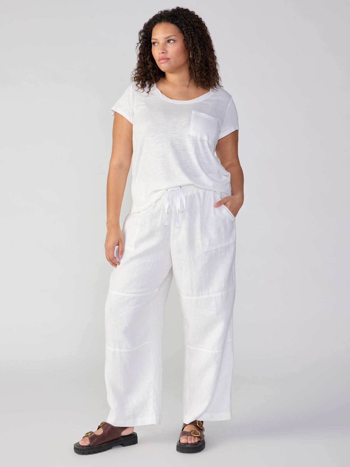 A person with curly hair models a relaxed, casual outfit consisting of the Traveler Twist Tee White from Sanctuary Clothing's Inclusive Collection and loose-fitting white drawstring pants. They are also wearing dark brown sandals and standing against a plain background.