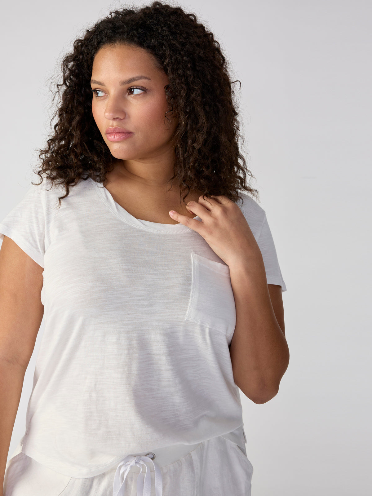 A person with curly hair is looking to the side, wearing the Traveler Twist Tee White from the Inclusive Collection by Sanctuary Clothing. The t-shirt features short sleeves and a pocket on the left side. The individual is holding a piece of their hair with one hand against a plain, light-colored background.