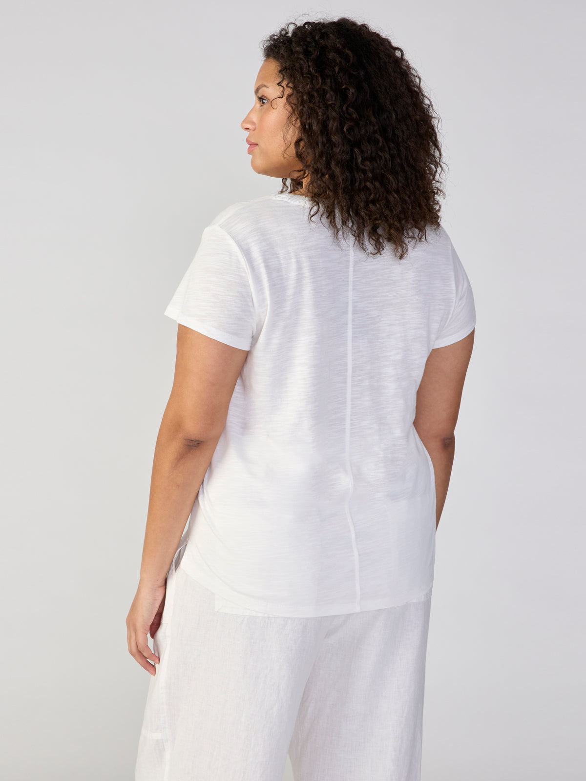 A woman with curly hair is turned slightly to her right, facing away from the camera. She is wearing Sanctuary Clothing's Traveler Twist Tee White from their Inclusive Collection, paired with white pants, against a light gray background.