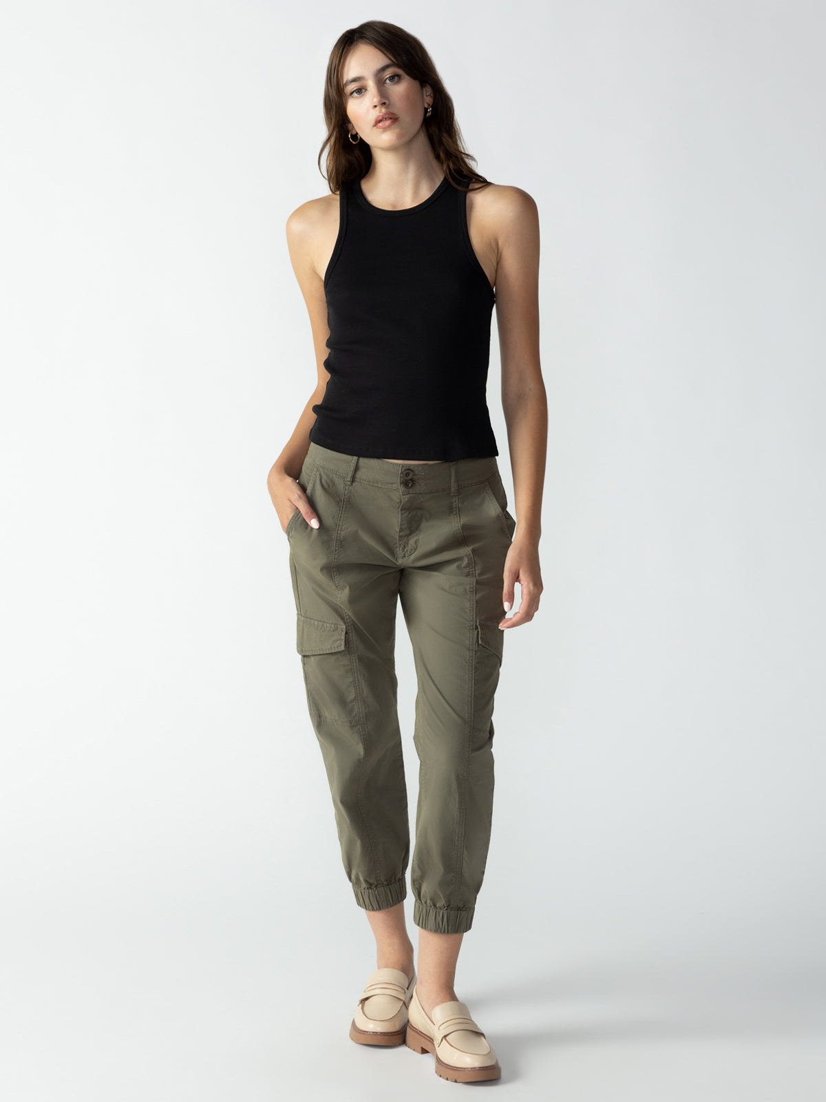 A woman stands against a plain background, wearing a Ribbed Tank Top Black by Sanctuary Clothing and olive green cargo pants with gathered ankles. She has one hand in her pocket and the other by her side. She is also wearing beige loafers and has long brown hair.