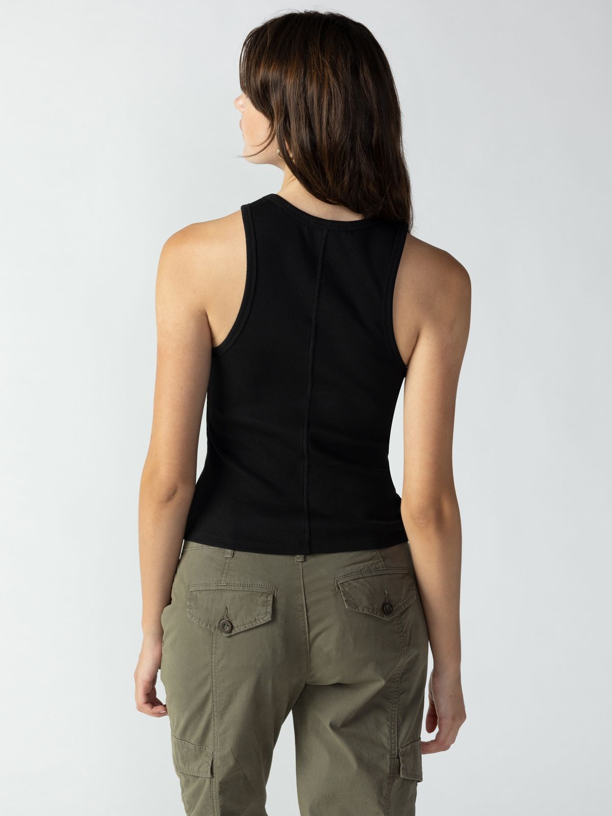 A person with dark hair is facing away from the camera, wearing a Ribbed Tank Top Black by Sanctuary Clothing and olive green cargo pants. The background is plain and light-colored.