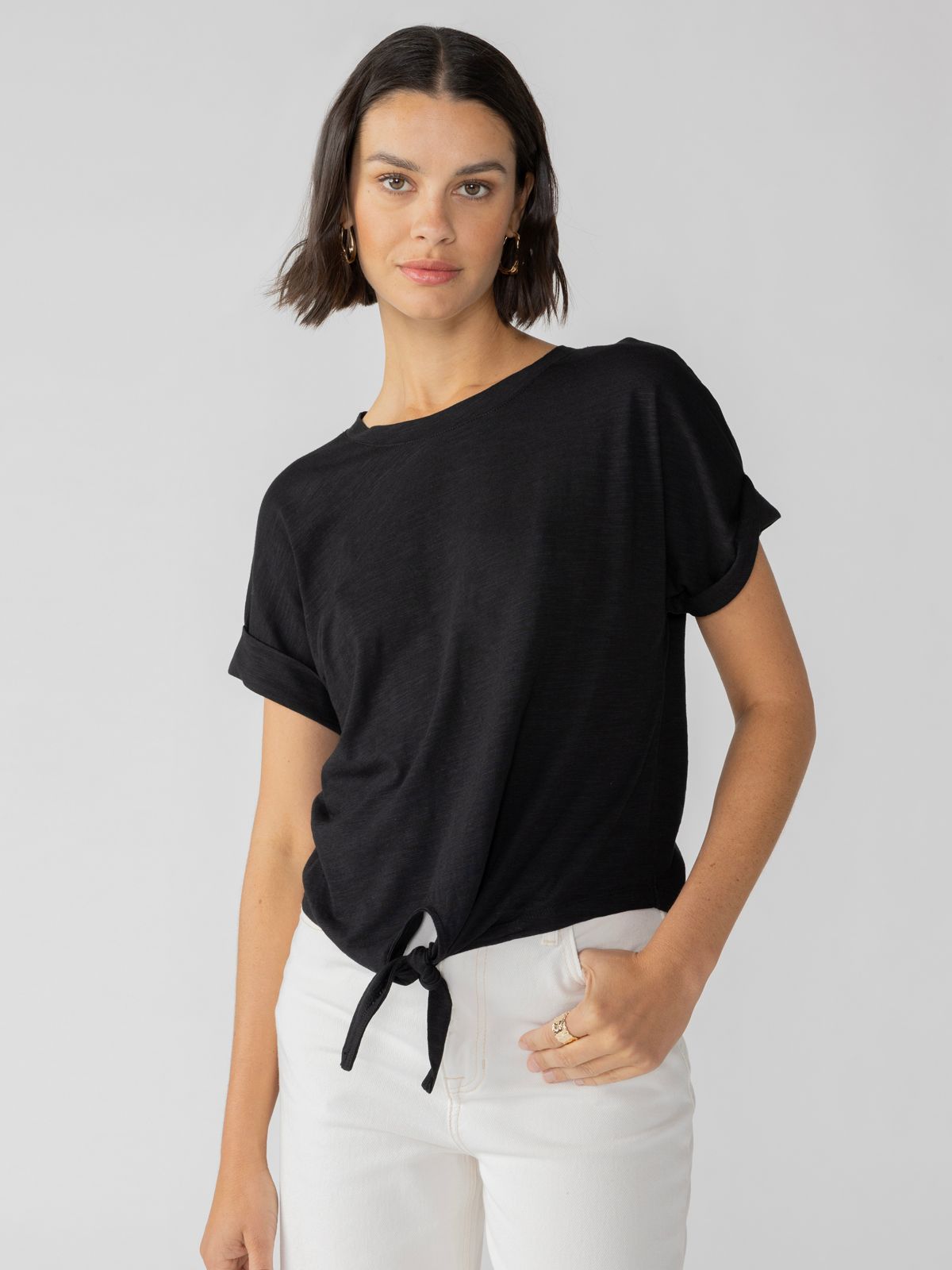 A person with shoulder-length brown hair is wearing the All Day Tie Tee Black from the Inclusive Collection by Sanctuary Clothing, paired with white pants. They are standing against a plain light gray background, with one hand in their pocket and a relaxed expression on their face.