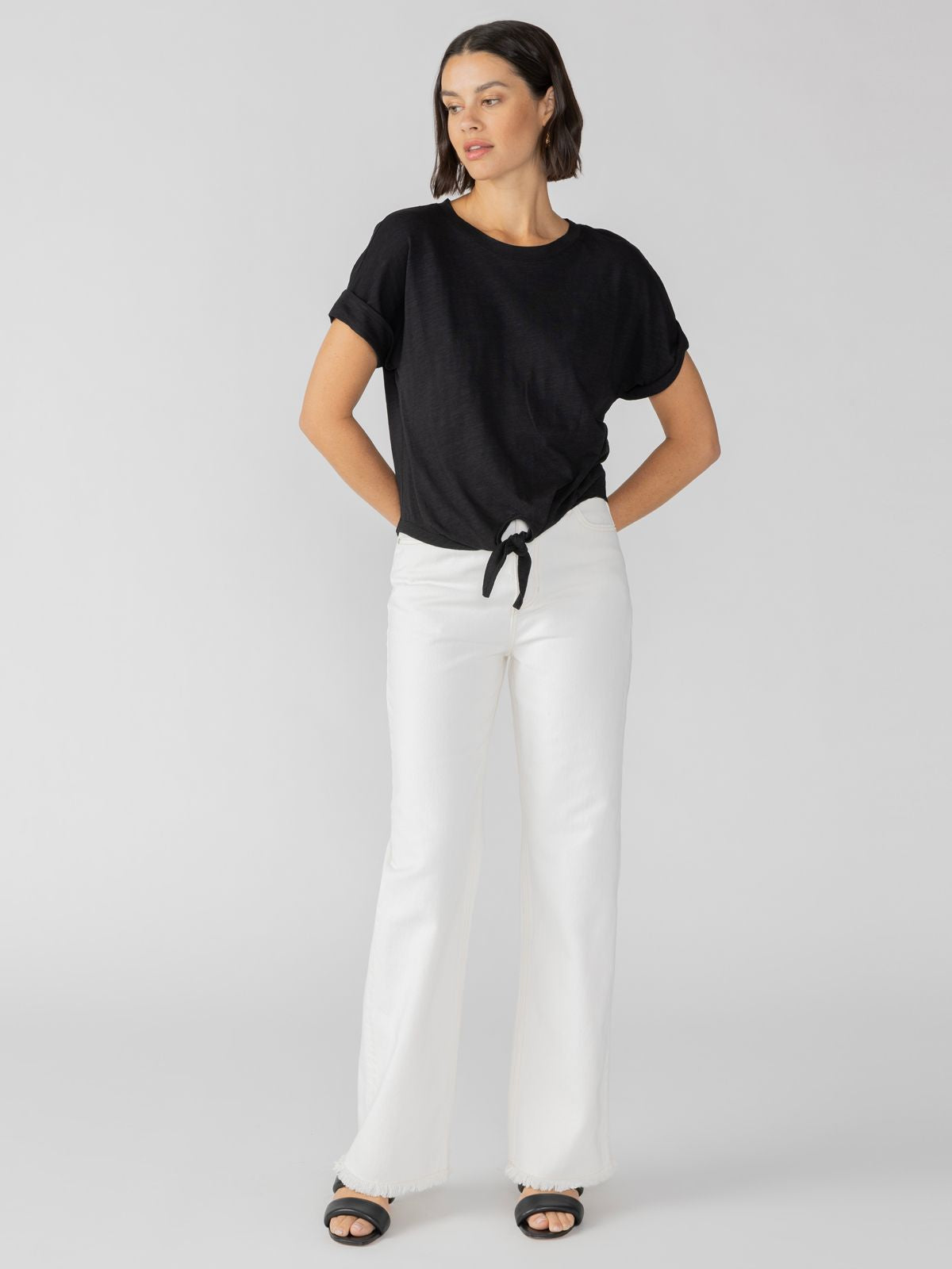 A person with shoulder-length hair wearing the All Day Tie Tee Black Inclusive Collection by Sanctuary Clothing, paired with white high-waisted pants and black open-toe sandals, stands against a plain gray background. Their hands are tucked into their pockets, and they are looking slightly to the side.