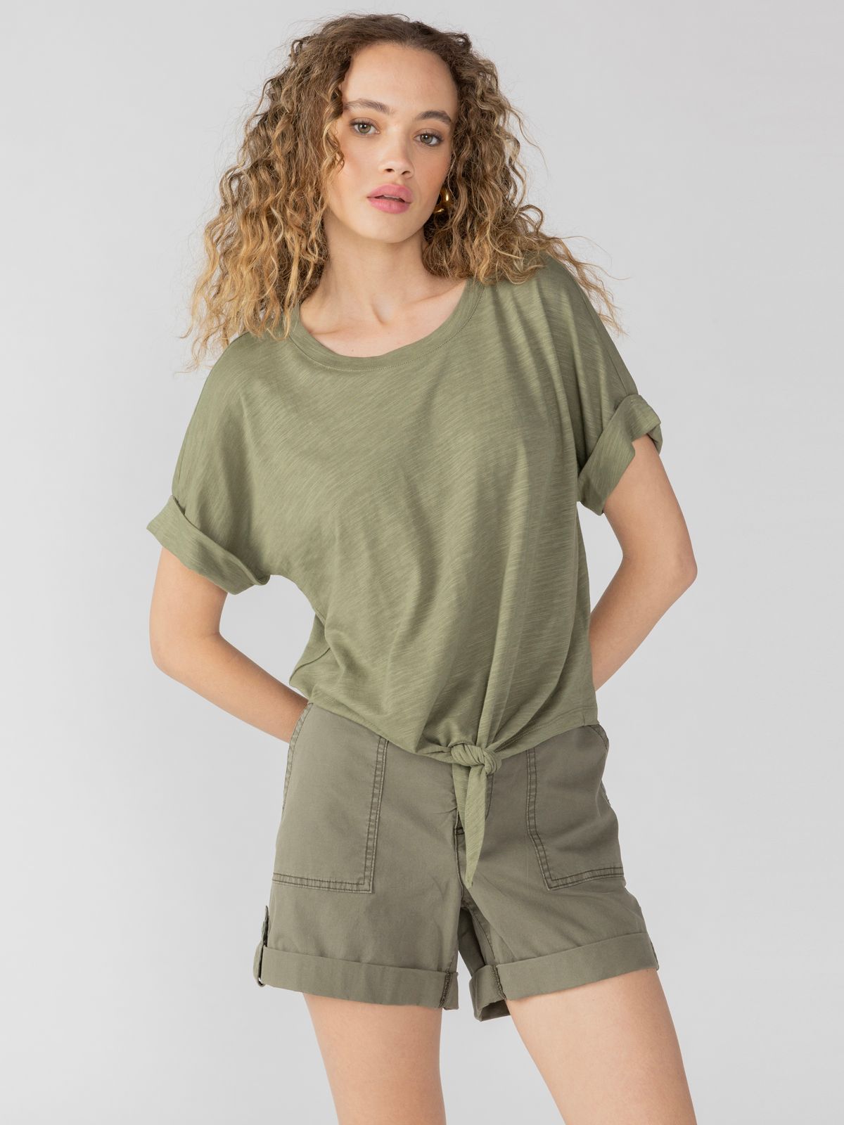 A person with curly hair is wearing the All Day Tie Tee from Sanctuary Clothing in Trail Green and matching green shorts. They're posing against a plain, light gray background.