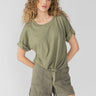 A person with curly hair is wearing the All Day Tie Tee from Sanctuary Clothing in Trail Green and matching green shorts. They're posing against a plain, light gray background.