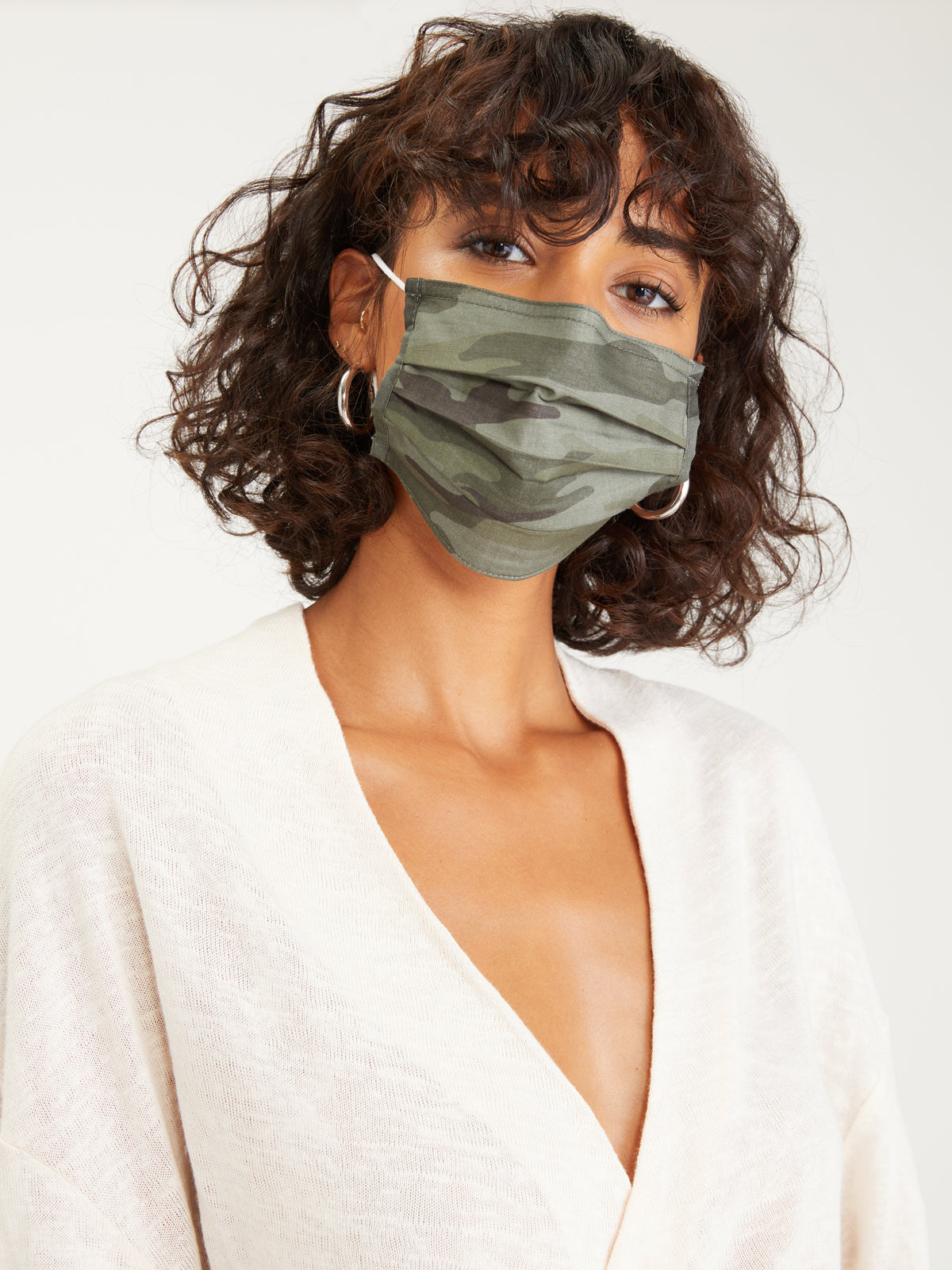 A person with wavy shoulder-length hair is wearing a Sanctuary Clothing Lifestyle Mask in a camouflage pattern and a light cream-colored V-neck top. They have hoop earrings and are looking directly at the camera, with a solid, light-colored background behind them.