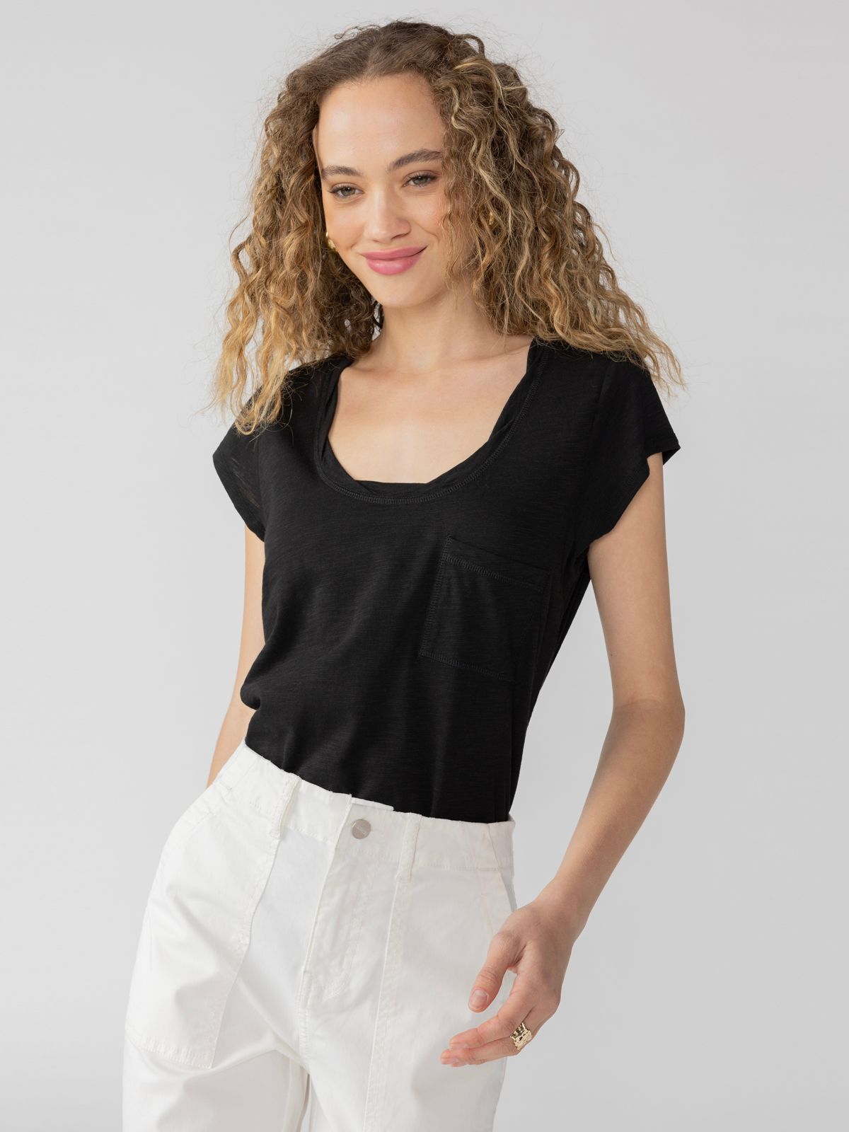 A person with wavy hair smiles while standing against a plain background. They are wearing the Traveler Twist Tee Black from Sanctuary Clothing and white high-waisted pants. Their left hand is slightly raised, and they have a relaxed, casual pose.