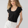 A person with wavy hair smiles while standing against a plain background. They are wearing the Traveler Twist Tee Black from Sanctuary Clothing and white high-waisted pants. Their left hand is slightly raised, and they have a relaxed, casual pose.