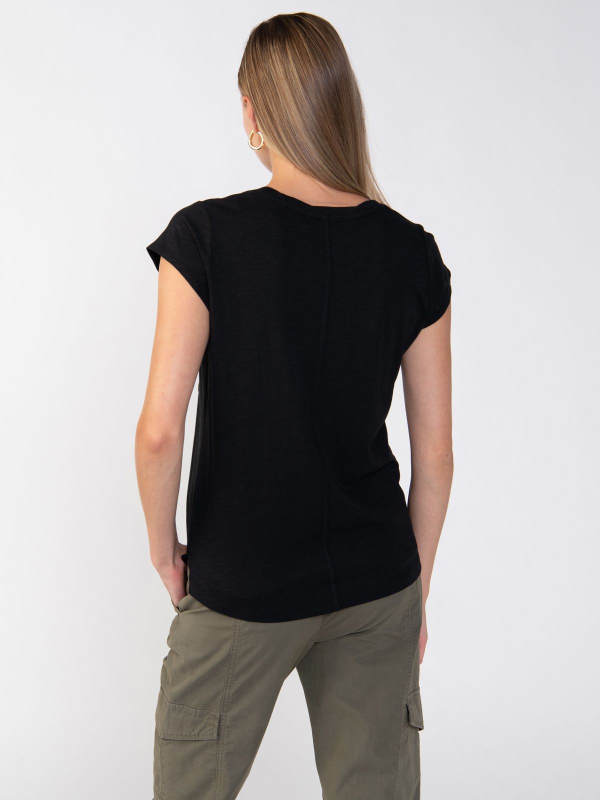 A person with long, straight hair is standing with their back to the camera, wearing a Sanctuary Clothing Traveler Twist Tee in black and olive green cargo pants. The background is plain white.