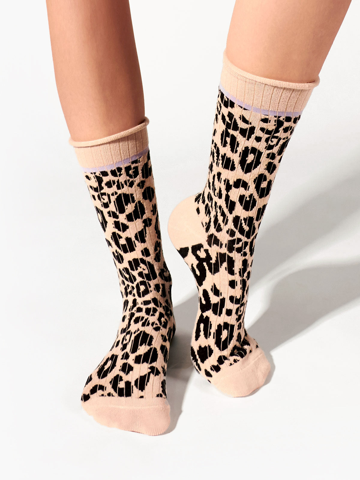 Two legs are adorned with a pair of Classy Cat Socks from Sanctuary Clothing, featuring a beige base with a striking black leopard print design. The light pink cuffs, heels, and toes offer a delightful contrast to the animal print pattern. The background is a simple, light-colored surface.