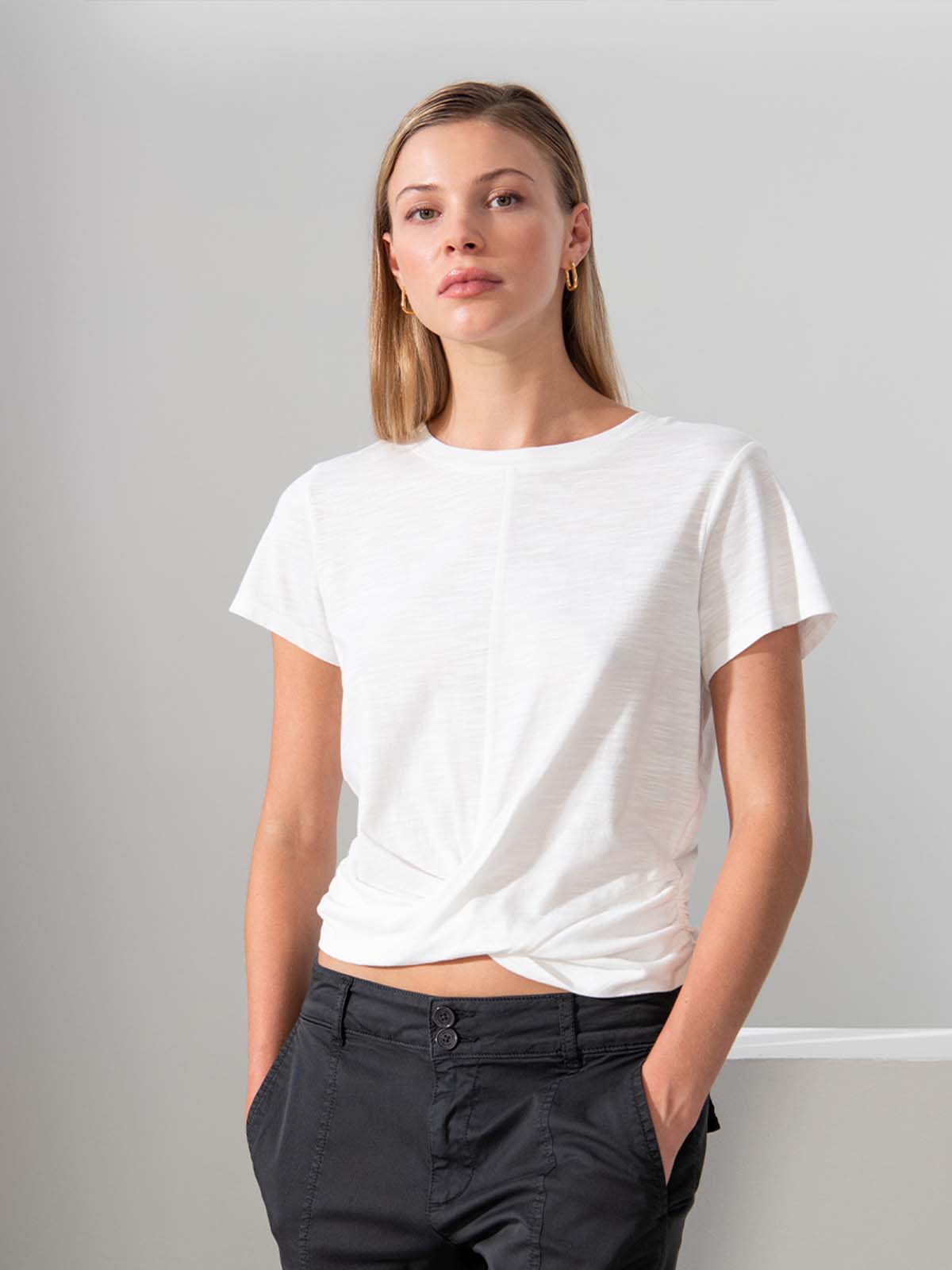 A woman with long blonde hair is standing against a simple background, wearing the Highstreet Tee White by Sanctuary Clothing, which features a twist detail at the front, paired with black pants. She has a serious expression and is looking directly at the camera. She is also wearing small hoop earrings.