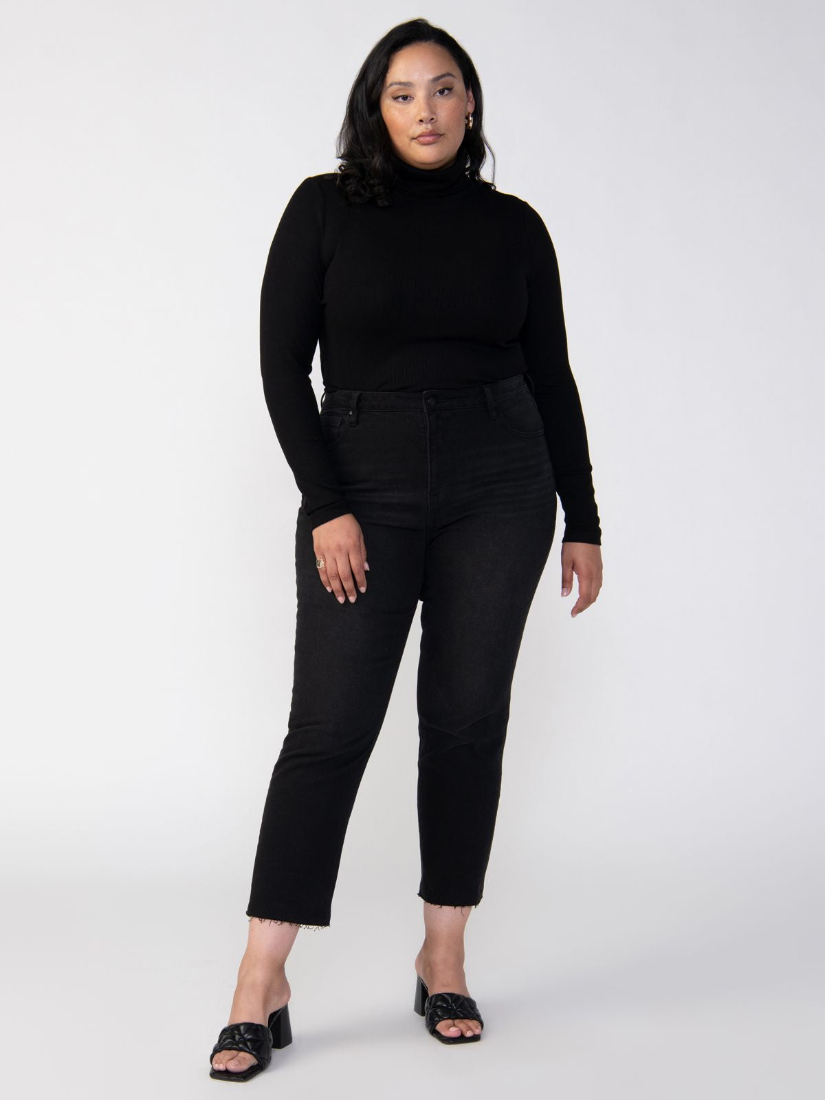 A person with medium-length dark hair is standing against a plain white background, wearing the Essential Turtleneck in black from Sanctuary Clothing's Inclusive Collection, paired with high-waisted black jeans and black open-toed shoes.