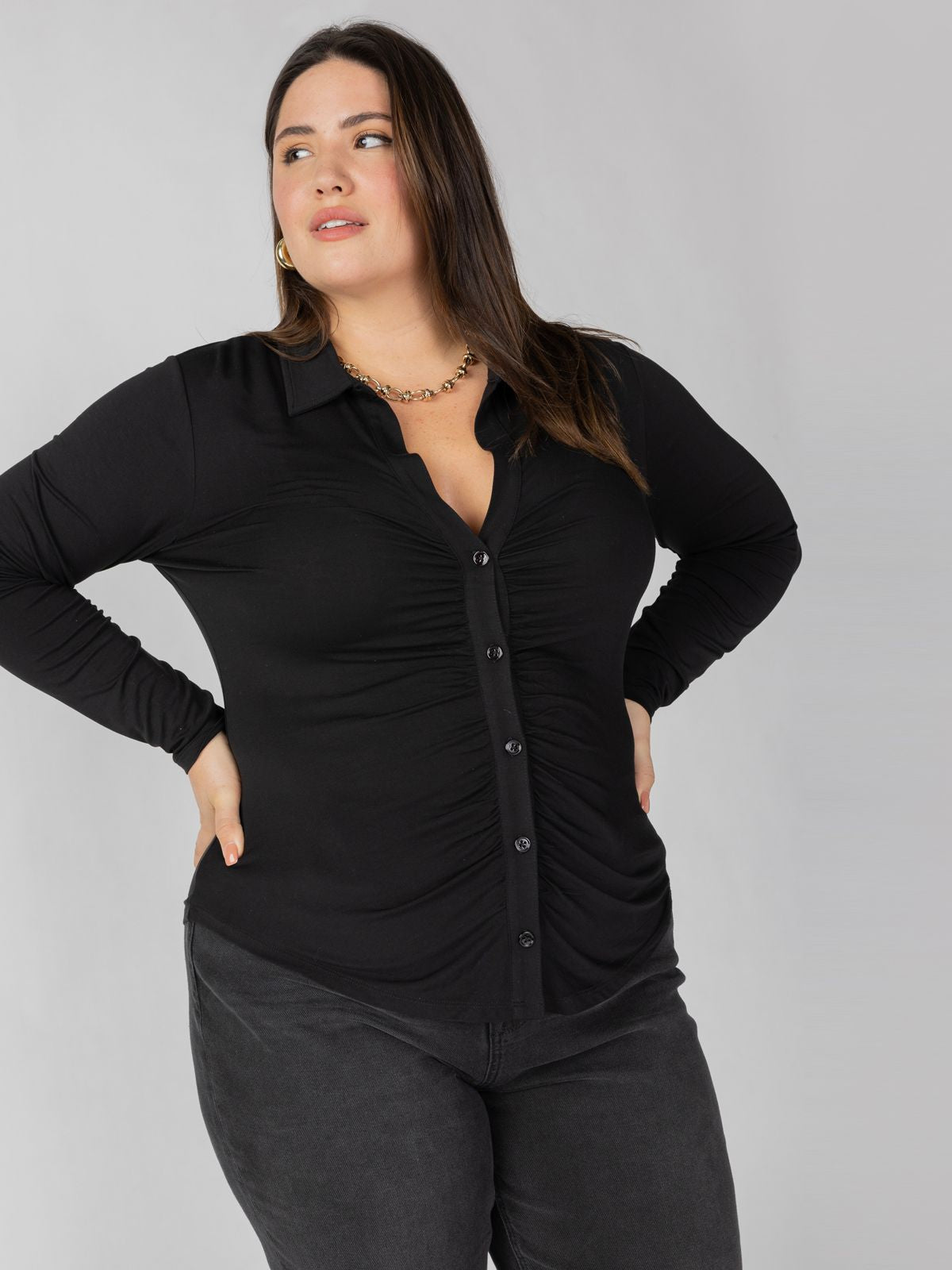 A person with long dark hair stands against a plain background, wearing the Dreamgirl Button Up Black Inclusive Collection shirt by Sanctuary Clothing, which features ruching down the center, paired with dark pants. They have their hands on their hips and are looking slightly to the side with a neutral expression.