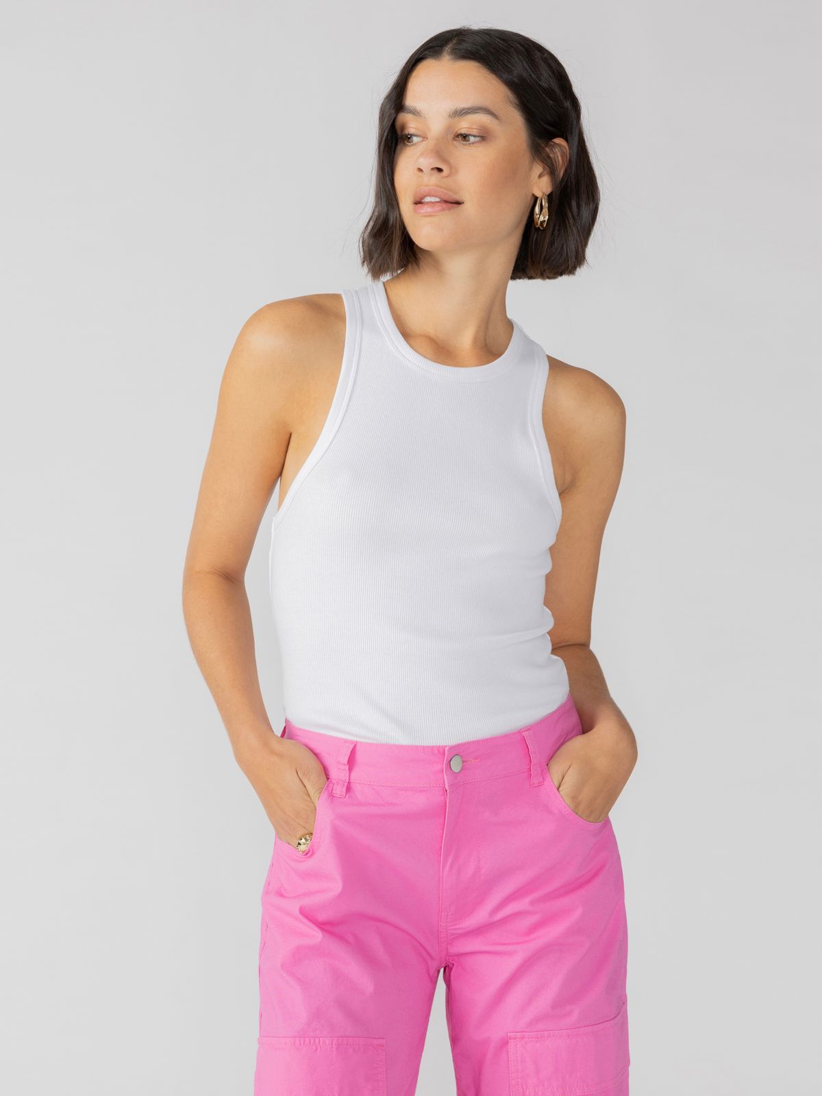 A woman with a bob haircut is wearing a Ribbed Tank Top in White from Sanctuary Clothing and bright pink pants. She has her hands in her pockets and is looking slightly to the side. The background is plain white.